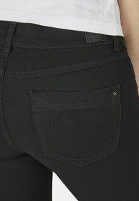 Svarta jeans med en slät yta, försedd med bakfickor, vertikala sömmar och kontrasterande sömnadsdetaljer. Minimalistisk design utan utsmyckningar.