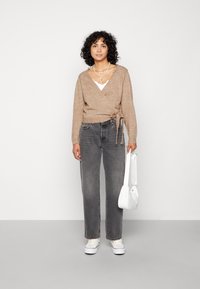 Brun inslagsko med en knytning, grå raka jeans och vita sneakers. Modellen håller en liten vit väska och är stylad med lager-på-lager halsband.