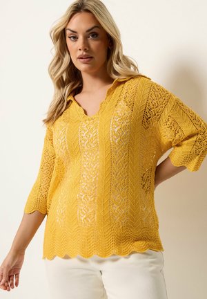 Blonde Frau trägt einen gelben Spitzenpullover mit gewellten Kanten und dreiviertellangen Ärmeln, kombiniert mit weißen Hosen, posiert mit einer Hand in der Hüfte.