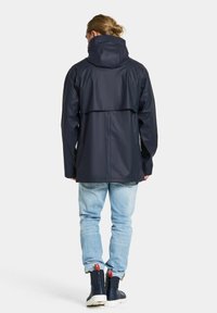 Marinblå vattentät jacka med hög krage, ryggventilation och justerbar huva. Kombinerad med ljusblå jeans och marinblå stövlar.