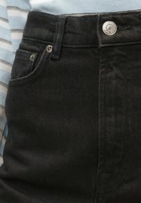 Μαύρα τζιν παντελόνια με κλασικό σχέδιο πέντε τσεπών, ασήμι κουμπί και υποτονική υφή, συμπληρωμένα από μια ριγέ μπλούζα στο φόντο.