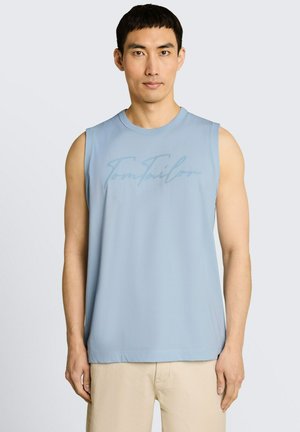 Man draagt een lichtblauw mouwloos topje met subtiel "Tom Tailor" logo en beige broek, staand tegen een eenvoudige lichte achtergrond.