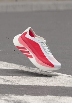 Κόκκινο και λευκό παπούτσι τρεξίματος στον αέρα πάνω από άσφαλτο με λευκή ριγωτή διάβαση πεζών, μικρά θραύσματα ορατά κάτω από την σόλα.