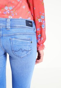 Světle modré džíny s úzkým střihem, které mají design zadní kapsy se stehovými akcenty a značkovou nášivkou na pase.