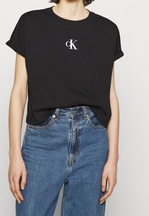 Person trägt ein schwarzes T-Shirt mit kurzen Ärmeln und weißem "cK"-Logo, das in eine hoch taillierte blaue Jeans gesteckt ist.