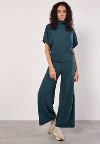 Zelenomodrá pletená souprava se zkrouceným vysokým výstřihem a širokými kalhotami. Materiál má jemnou texturu; outfit je doplněn neutrálními teniskami.