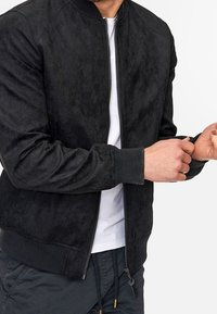 Schwarze Wildleder-Bomberjacke mit gerippten Bündchen und Kragen, ausgestattet mit einem Frontreißverschluss und glatter Textur. Wird über einem weißen T-Shirt getragen.