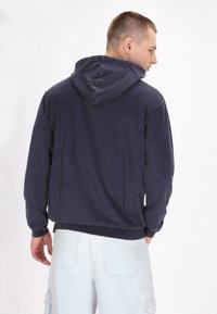 Felpa blu navy con cappuccio regolabile, polsini e orlo a coste, indossata sopra pantaloni cargo in denim chiaro, che mostrano un design e una texture casual.