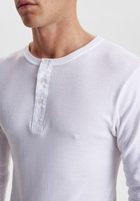 Hvit langermet henley-skjorte laget av bomull, med rund hals, tre knappelukking og ribbestrikket krage og mansjetter.