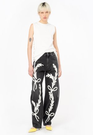 Top senza maniche bianco, jeans neri a gamba larga con motivo floreale ricamato bianco e fiocchi, e ballerine gialle a punta su uno sfondo semplice.