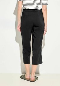 Schwarze cropped Hosen mit einem elastischen Bund, ausgestattet mit zwei Gesäßtaschen und einer schlanken, glatten Textur. Getragen mit lässigen Sandalen.