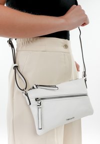Borsa a tracolla bianca in pelle con due scomparti con zip, dettagli neri e tracolla regolabile, esposta su uno sfondo chiaro.