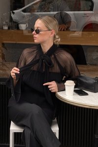 Blusa nera a pieghe con scollatura annodata, maniche lunghe e texture a strati. Abbinata a pantaloni neri, una borsa piccola e una tazza di caffè su un tavolo.
