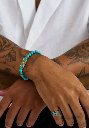 Turkis perlearmbånd med en dekorativ midterdel med et øje-design, båret på et tatoveret håndled mod en ensfarvet baggrund.
