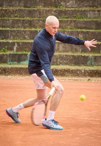 Joueur de tennis portant une veste marine et un short clair, utilisant une raquette pour frapper une balle jaune sur un court en terre battue orange, avec des chaussures bleues aux accents roses.