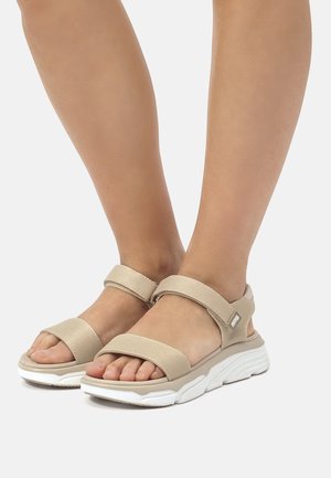 Pieds portant des sandales beiges ouvertes à l'avant avec des brides Velcro et une semelle blanche épaisse sur un fond clair uni.