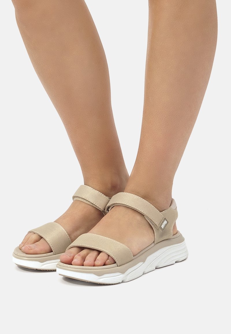 Pieds portant des sandales beiges ouvertes à l'avant avec des brides Velcro et une semelle blanche épaisse sur un fond clair uni.