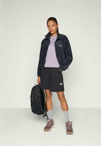 Young woman with braided hair wearing a black jacket, purple shirt, black shorts, gray socks, and purple sneakers, holding a black backpack.