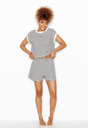 Giovane donna con capelli ricci che indossa una canotta a righe bianco e nero e pantaloncini abbinati, sorridente e che si sistema la camicia.