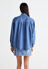 Blue denim shirt with long sleeves, button cuffs, a collar, pleated back, and a curved hem, worn over a lighter denim skirt.
