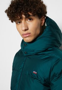 Tirkizna puffer jakna s visokim kragnom, dugmadi na pritisak i malim Levi's logotipom na prsima. Glatka, sjajna tkanina s kviltanom teksturom.