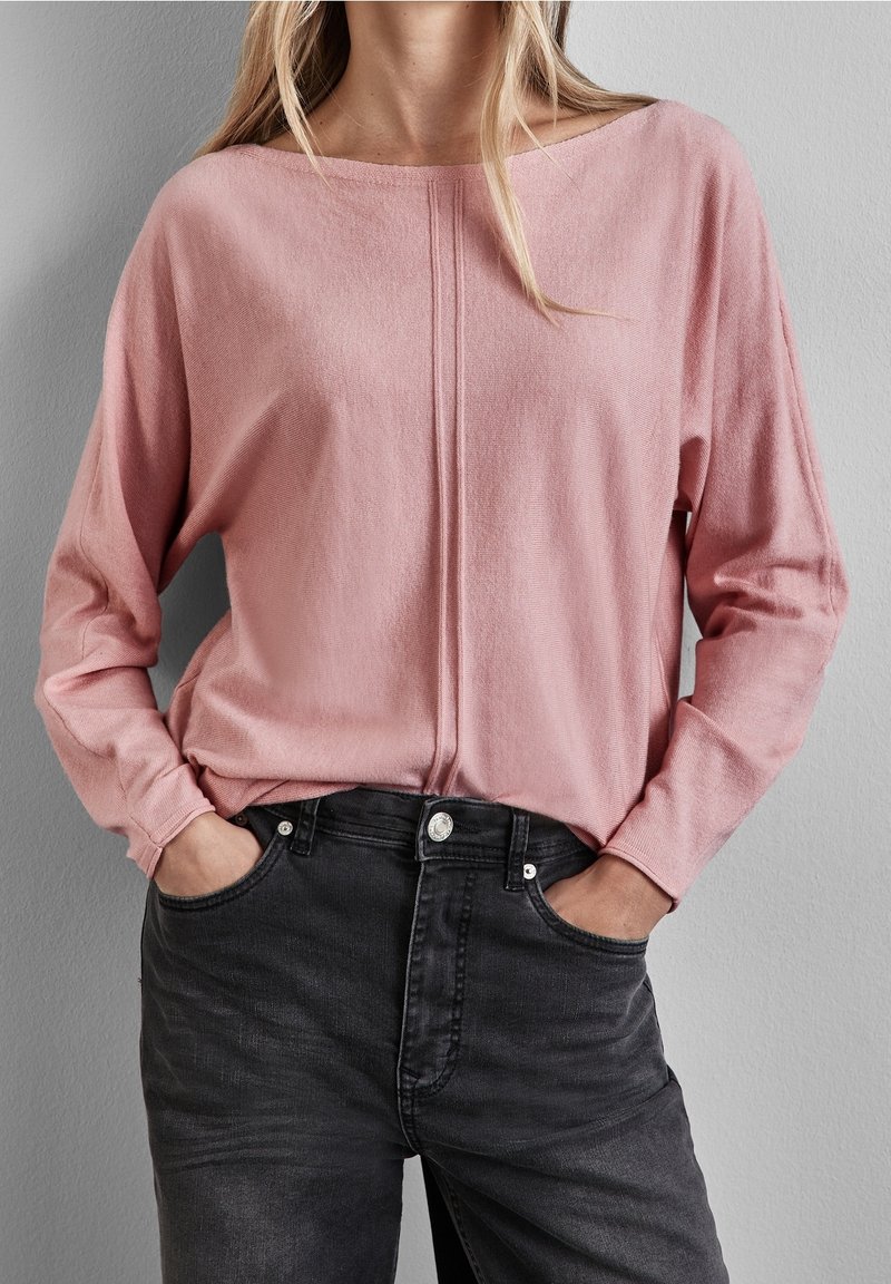Haut rose clair à manches longues avec un col bateau et une couture centrale ; porté avec un jean noir taille haute, avec des poches avant et une fermeture à bouton.