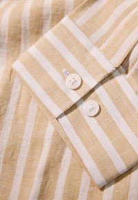 Camicia a maniche corte con un motivo a righe gialle e bianche, dotata di colletto e due bottoni tondi sulla patta. Materiale in lino.