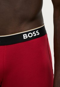 Gros plan sur la taille d'une personne portant un sous-vêtement rouge avec une ceinture noire étiquetée « "BOSS" » en lettres blanches.