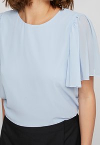 Blusa azzurro chiaro con maniche corte a svolazzare, scollatura rotonda e una texture di tessuto liscia. Abbinata a un pantalone nero aderente.