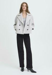 Jeune femme portant un manteau croisé gris clair, un haut gris, un pantalon noir et des bottes à talons pointues noires, se tenant devant un fond uni.