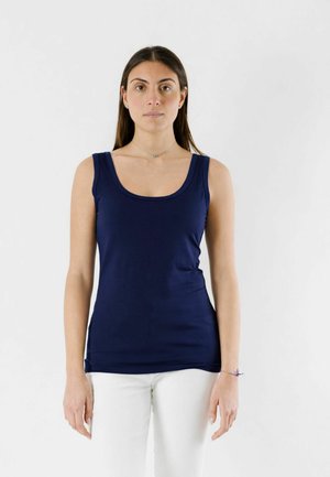 Donna con lunghi capelli scuri che indossa un top blu navy senza maniche e pantaloni bianchi, in piedi contro uno sfondo chiaro e semplice.