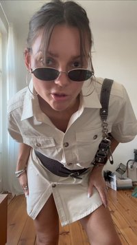 Femme portant une robe beige boutonnée avec une ceinture noire, de grandes boucles d'oreilles créoles et des lunettes de soleil foncées, se penchant légèrement en avant dans une pièce avec un sol en bois.