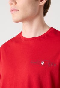 T-shirt en coton rouge avec un col rond, présentant un petit logo gris imprimé sur la poitrine. Texture lisse et coupe décontractée.