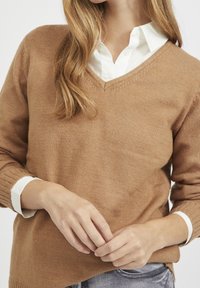 Bruine, gebreide V-hals trui met ribgebreide manchetten, gelaagd over een wit overhemd met kraag, met een zachte textuur en een ontspannen pasvorm.