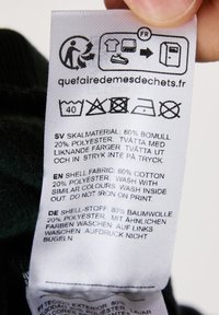 Etichetta di cura dei tessuti su un tessuto bianco con istruzioni multilingue. Materiale: 80% cotone, 20% poliestere. Le icone di cura includono linee guida per il lavaggio e l'asciugatura.