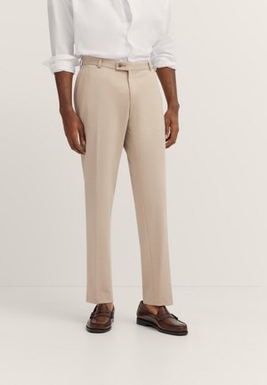 Mann trägt beige maßgeschneiderte Hose, weißes hochgekrempeltes Hemd mit langen Ärmeln und dunkelbraune Leder-Loafer, steht vor einfarbigem Hintergrund.