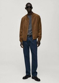 Giacca bomber in suede marrone sopra una camicia grigia, abbinata a jeans blu scuro e stivaletti neri, accessoriata con occhiali da sole neri.