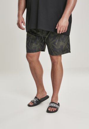 T-shirt oversize nera, pantaloncini da bagno verdi con motivo a palme e ciabatte nere con scritte bianche. Texture opaca e vestibilità casual.