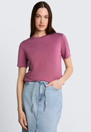 Femme aux longs cheveux bruns portant un t-shirt rose à manches courtes et une jupe en jean bleu clair taille haute avec ceinture à nouer, debout devant un fond uni.