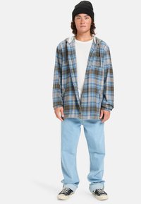 Grijs en blauw geruit flanellen shirt met een capuchon, gedragen over een wit t-shirt. Lichtblauwe jeans en zwarte sneakers maken de outfit compleet.