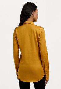 Camisa de seda amarela mostarda com mangas longas, colarinho pontudo e punhos com botões; textura suave e bainha arredondada.