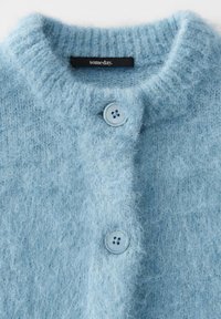 Cardigan morbido di un blu chiaro con due bottoni chiusi e un'etichetta nera con la scritta "someday" all'interno del colletto.