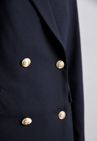 Gros plan d'un blazer croisé bleu marine foncé avec quatre boutons dorés embossés brillants à l'avant.