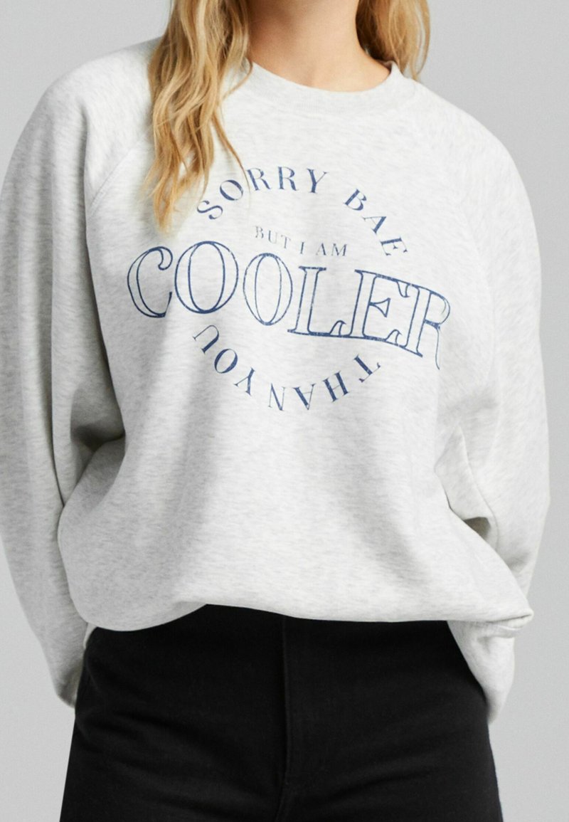 Femme portant un sweat-shirt gris clair avec un texte bleu disant "Sorry bae but I am cooler than you" et un pantalon noir sur un fond gris uni.