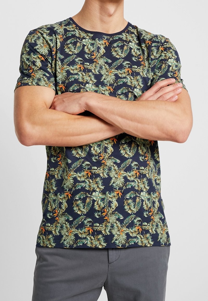 Mężczyzna w granatowej koszuli z krótkim rękawem w zielono-pomarańczowy tropikalny wzór, z założonymi rękami, w połączeniu z szarymi spodniami.