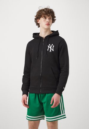 Felpa nera con zip e logo bianco di New York, abbinata a pantaloni corti in rete verdi con strisce bianche all'orlo. Tessuto morbido e vestibilità rilassata.