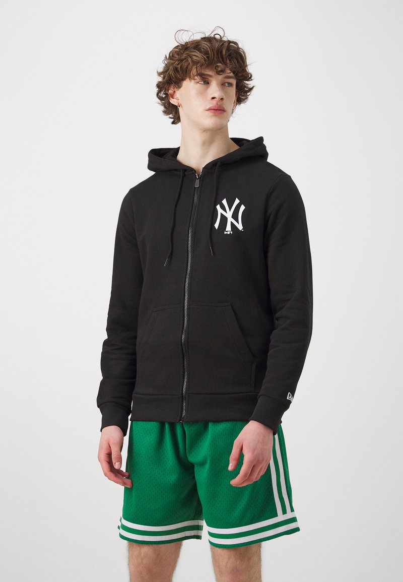 Čierna mikina s kapucňou na zips s bielym logom New York, skombinovaná so zelenými sieťovými šortkami s bielymi pruhmi na leme. Mäkká tkanina a uvoľnený strih.