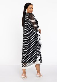 Schwarz-weißes Maxikleid mit Polka-Dots aus transparenter Stoff, das mit weißen Fransen entlang des Saums und der Ärmel verziert ist. Kombiniert mit weißen Sandalen.