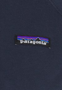 Tessuto blu scuro con una texture a coste caratterizzato da una rettangolare patch nera che mostra il logo "patagonia" in bianco con un grafico di montagne colorato.