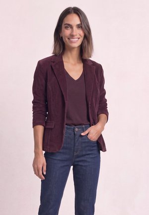 Femme souriante aux cheveux bruns mi-longs, portant un blazer violet en velours côtelé, un t-shirt violet foncé à col en V et un jean bleu avec une main dans la poche.